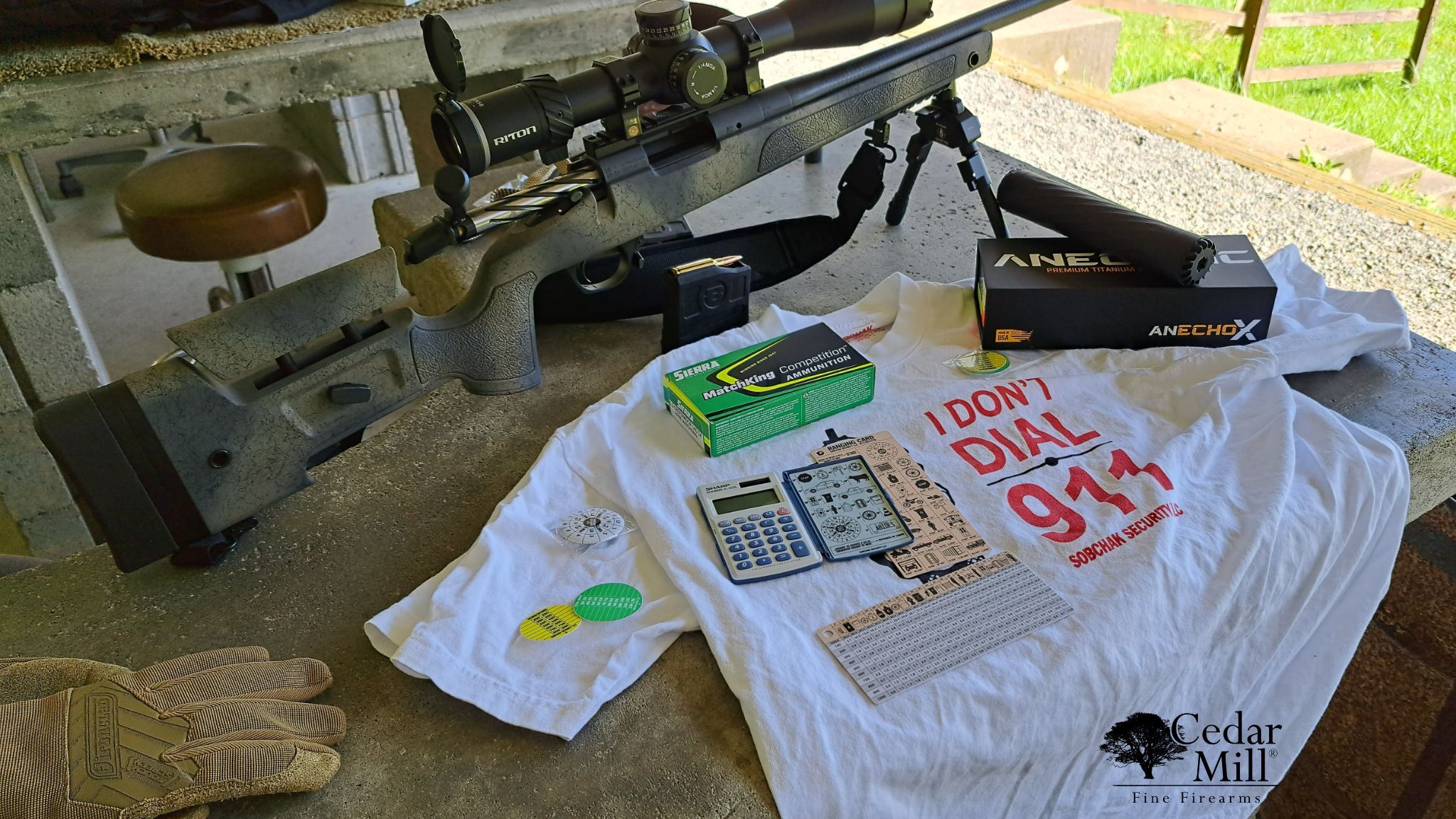 The “Obviously You’re Not a Golfer” MOA Pack from Sobchak Security is laid out on a shooting bench, featuring essential tools for precision shooting: MOA dope cards, ballistic calculator, range cards, and data wheels. The centerpiece is a bold white T-shirt with “I Don’t Dial 911 – Sobchak Security” in red text. Nearby are premium accessories including a precision rifle with a Riton optic, Sierra MatchKing competition ammo, gloves, and an AnechoX titanium suppressor, all staged at an outdoor range.